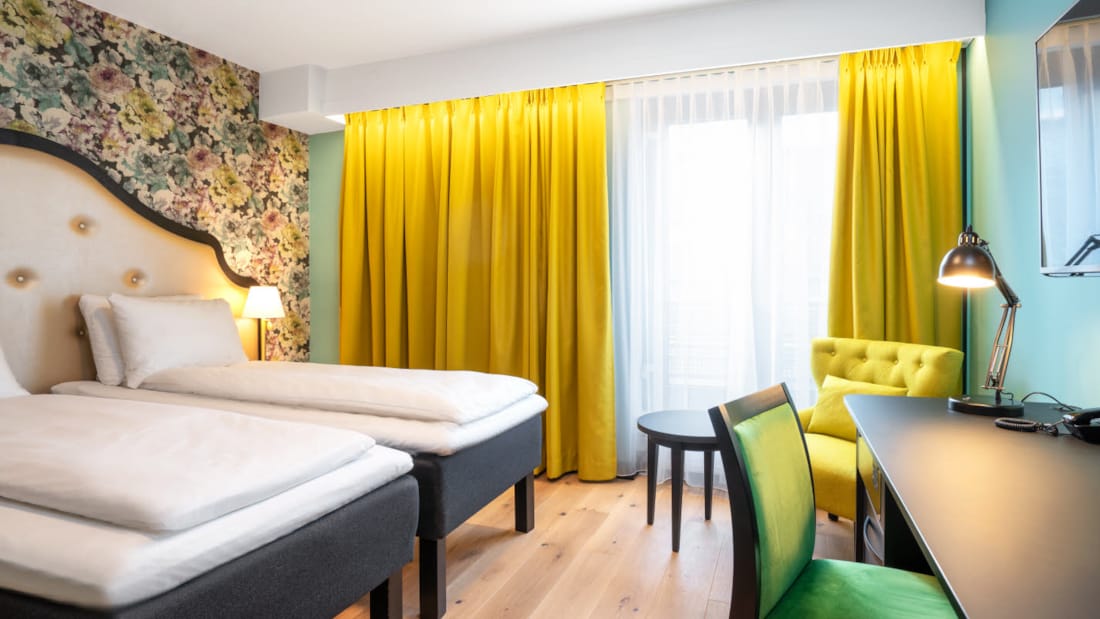 Två enkelsängar, blommig tapet bakom sängarna, gul stol, skrivbord och stora fönster med stora gula gardiner i Twin Room på Thon Hotel Cecil i centrala Oslo