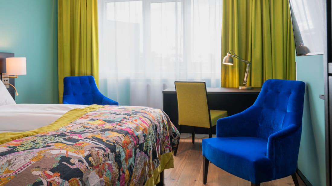 Twin room med hvidt sengetøj, mønstret sengetæppe, gule gardiner, skrivebord og blå lænestol på Thon Hotel Europa i Oslo centrum lige ved Slottsparken