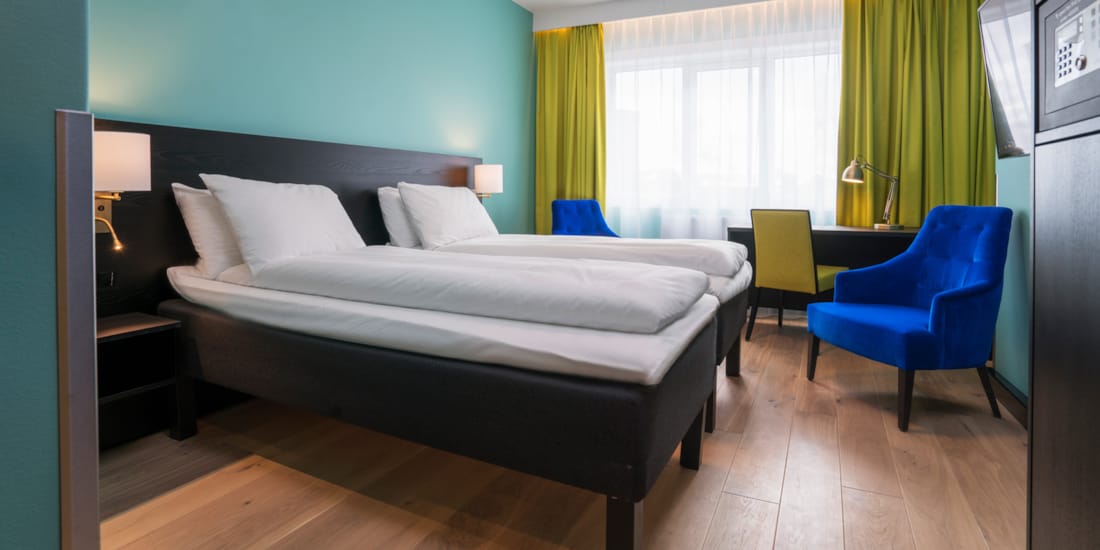 Twin room med hvit sengetøy, gule gardiner, skrivepult og blå lenestol på Thon Hotel Europa i Oslo sentrum rett ved Slottsparken