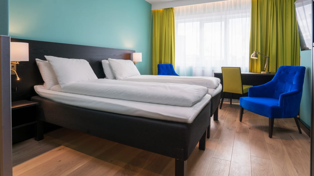 Twinrum med vita sängkläder, gula gardiner, skrivbord och blå fåtölj på Thon Hotel Europa i centrala Oslo nära Slottsparken