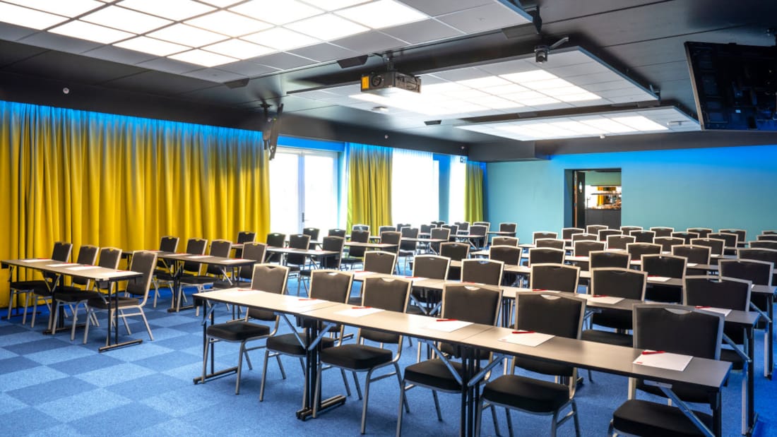 Konferenssal Bjerke med klassrumsmöblering med plats för 100 personer. Blå matta, gula gardiner och projektor på Thon Hotel Linne i Oslo