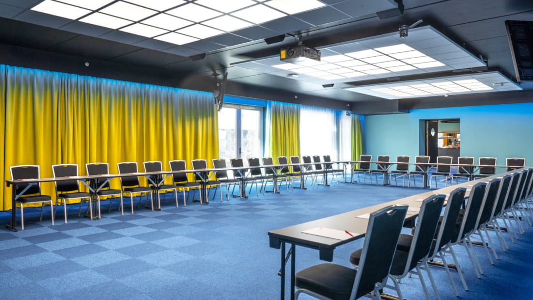 Konferenssal Bjerke med hästskomöblering med plats för 52 personer. Blå matta, gula gardiner och projektor på Thon Hotel Linne i Oslo