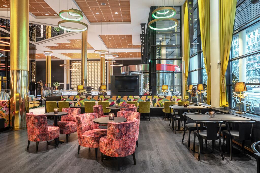 En restaurant med høy takhøyde, kunst på veggen, røde lenestoler og en bar med barkrakker.