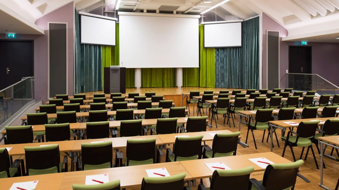 Grote vergaderruimte in klaslokaalopstelling met drie projectoren en podium