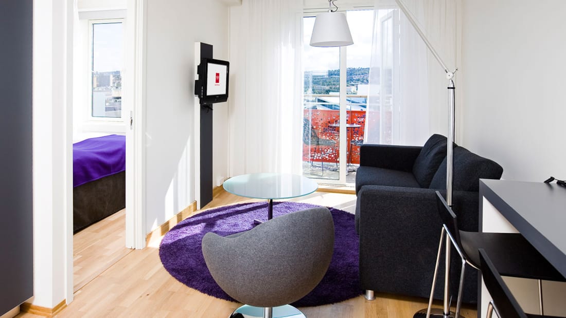 standard room triplefamily familieværelse stuesektion med sofa, sofabord, lænestol, gulvlampe, parketgulv, væghængt tv, balkon med udsigt over Oslo centrum, skrivebord og stole