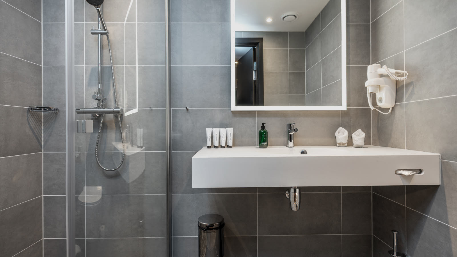 Hotellbaderom med dusj på venstre side og en hvit vask med et stort speil over, grå fliser på veggene. 