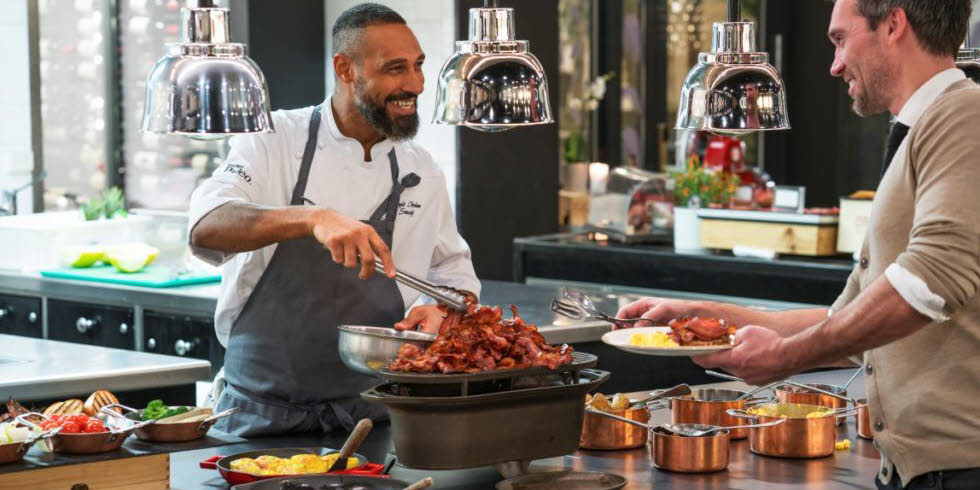 A happy breakfast chef serves a smiling guest bacon at Thon Hotel Rosenkrantz in Oslo city center.