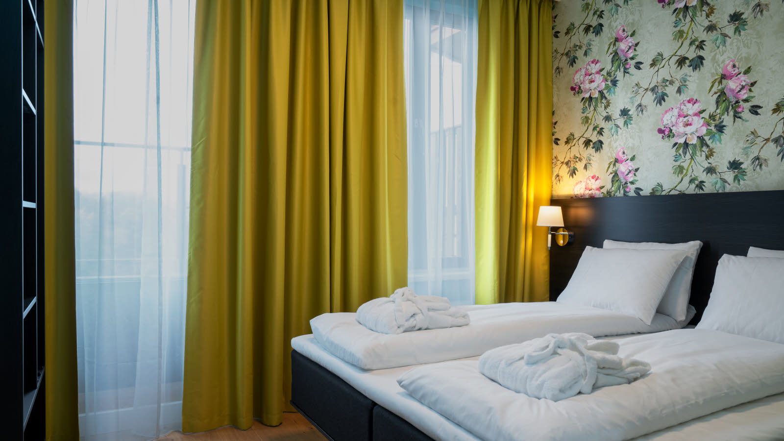 Hotel room with double bed. Made bed with duvets in white with towels on the bed, yellow curtains covering a large window and floral wallpaper in light green and pink behind the bed.