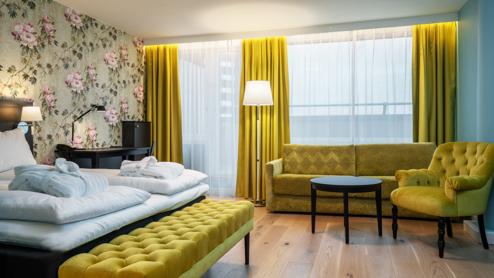 Large hotel room or small suite with yellow curtains, yellow bench in front of the bed and yellow armchair against green walls and patterned wallpaper behind the bed. Double bed with made bed and a large window.