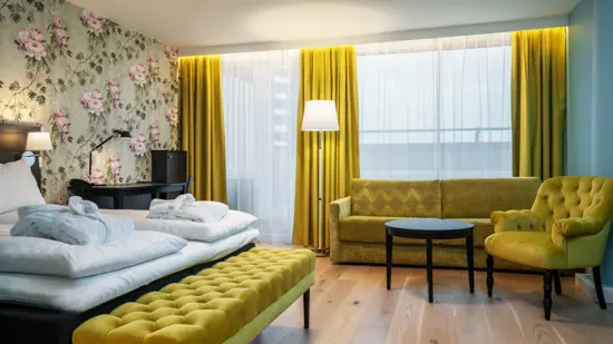 Stort hotellrom eller en liten suite med gule gardiner, gul benk foran sengen og en gul lenestol inn mot grønne vegger og mønstrete tapet bak sengen. Dobbeltseng med oppredd sengetøy og et stort vindu.