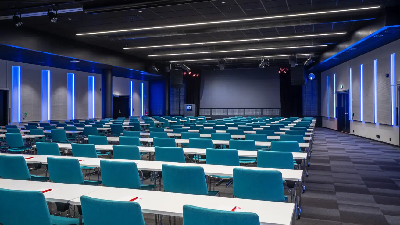 Large room seen from the rear in classroom layout, blue lighting on walls. Carpeted floor.
