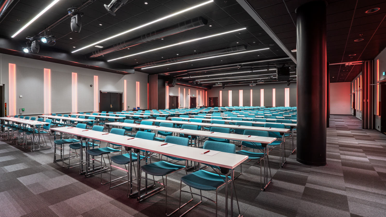 Storosalen hos Thon Hotel Storo sett forfra, rød belysning, klasseromsoppsett med langbord og 6 turkise stoler på ved hvert bord, skrivesaker på hver plass, teppebelagt gulv.