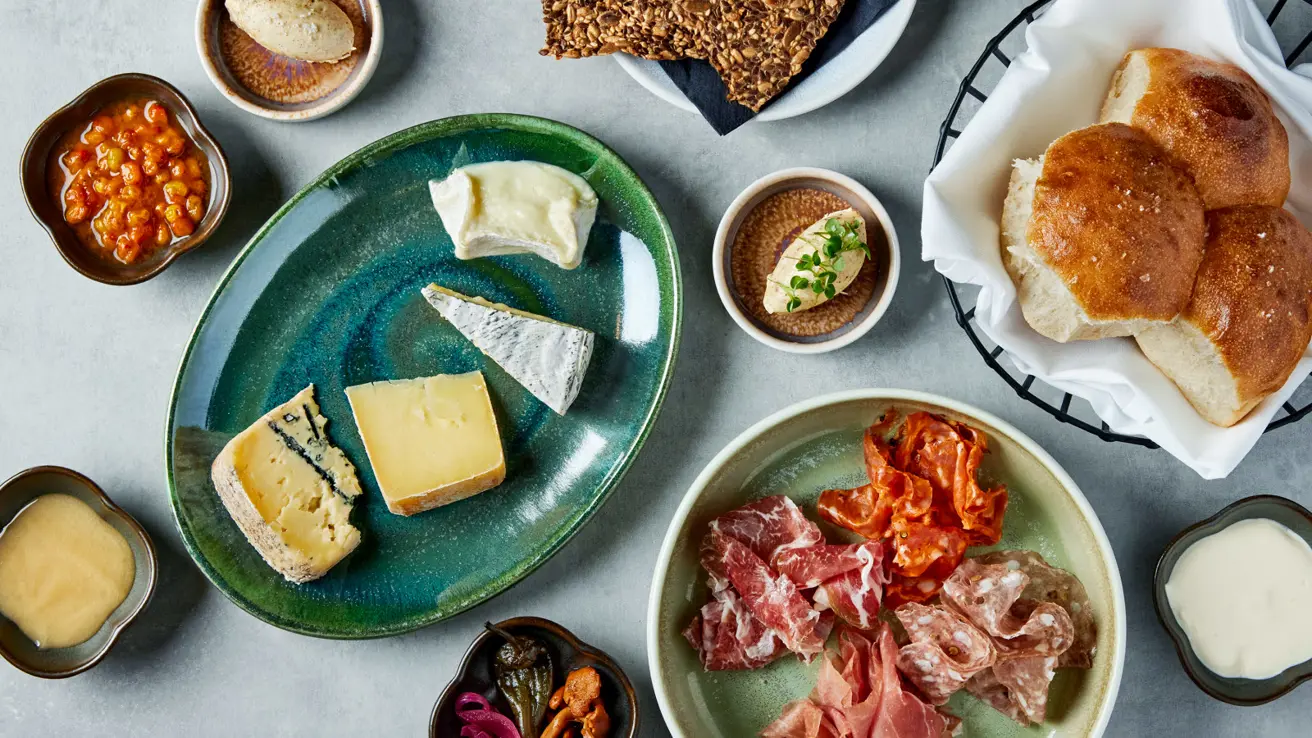 Utsnitt av flere oster og skinker på tallerkener på et bord med lys duk, sett ovenfra.