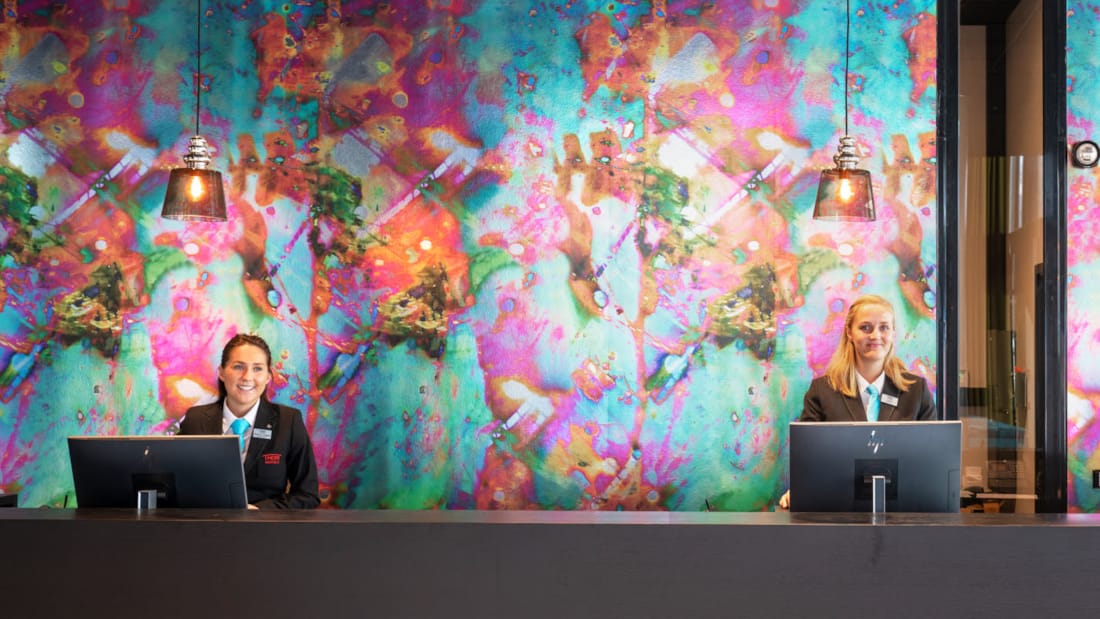 Two receptionists standing behind reception desk at Thon Hotel Storo. In the background is a colourful wall.