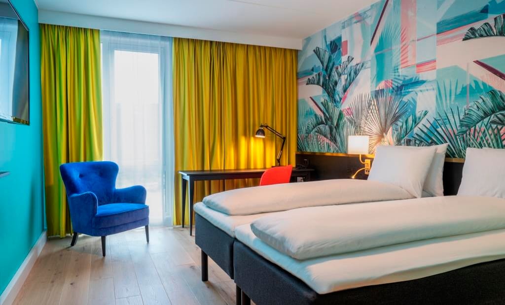 Del av dubbelsäng, arbetsbord och stor blå fåtölj, stora ljusa fönster och gula gardiner i dubbelrum på Thon Hotel Storo i Oslo
