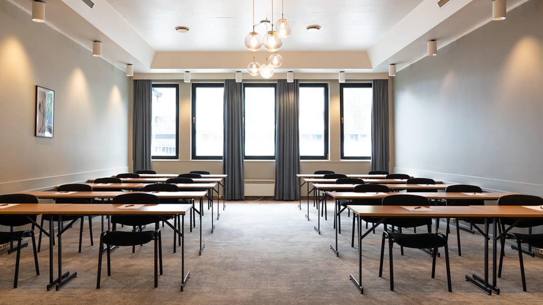 Lyst hvidt konferencelokale med to rækker stole og borde, højt til loftet og store lyse vinduer.
