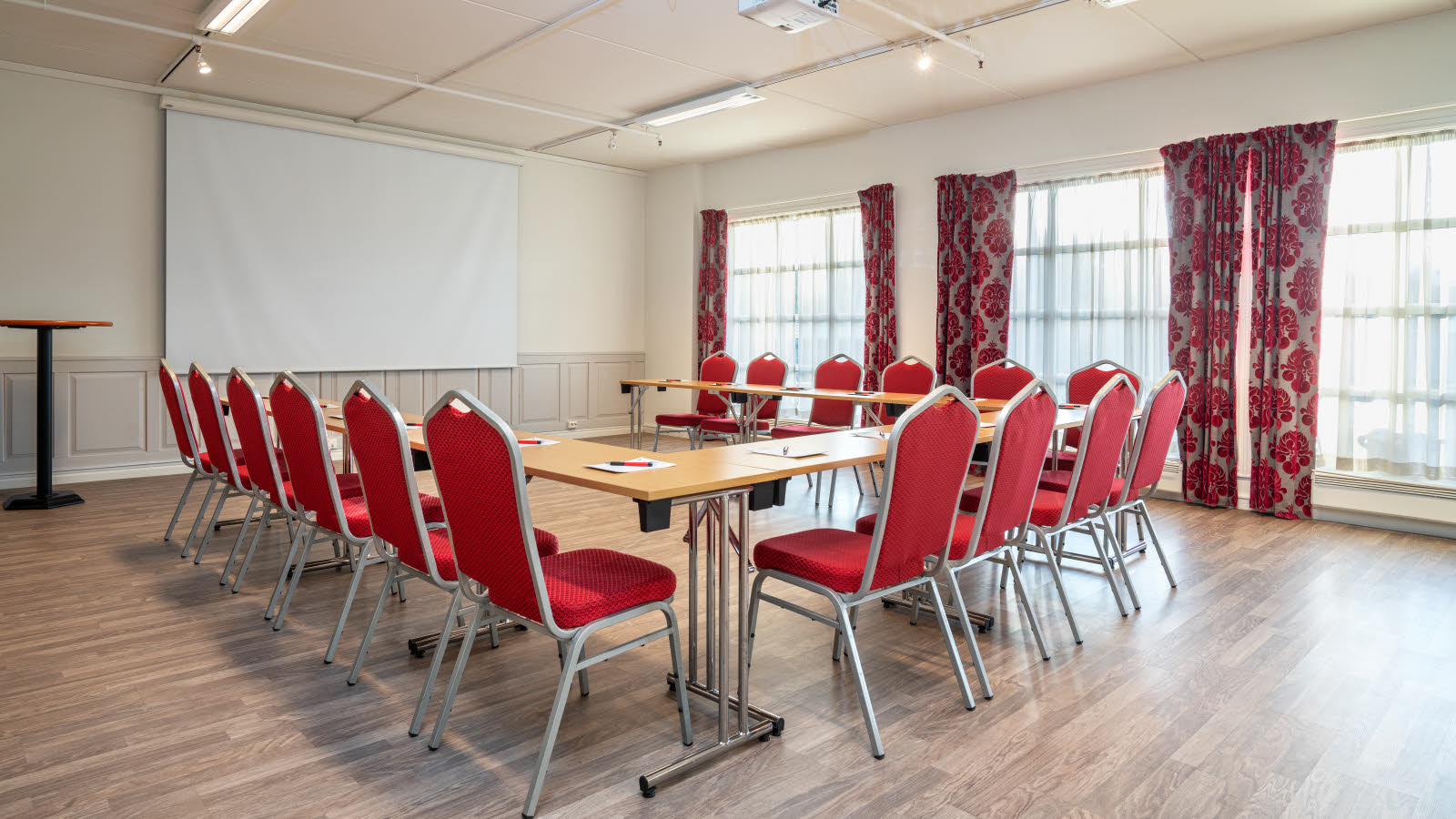 Stort og lyst møterom med vinduer på høyre side. Bord og røde stoler satt opp i U-form.