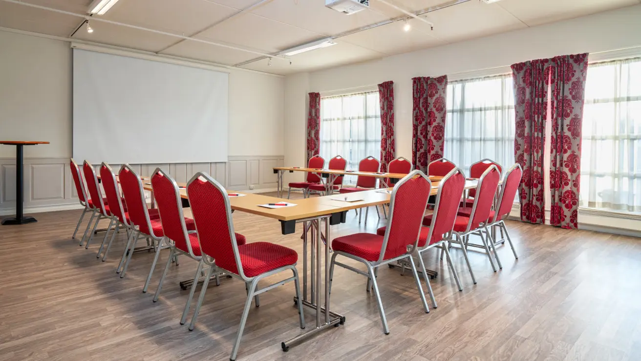 Stort og lyst møterom med vinduer på høyre side. Bord og røde stoler satt opp i U-form.