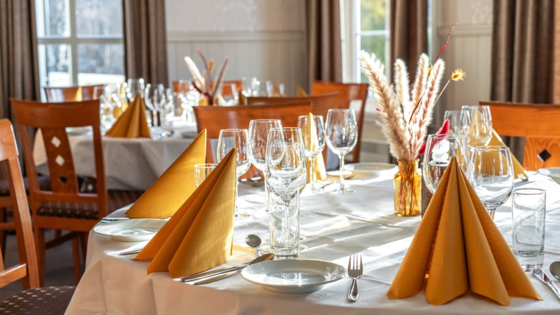 Utsnitt av to runde bord med hvite duker. Dekket med tallerkener, glass og gule servietter. Eikefargede stoler rundt bordene. 