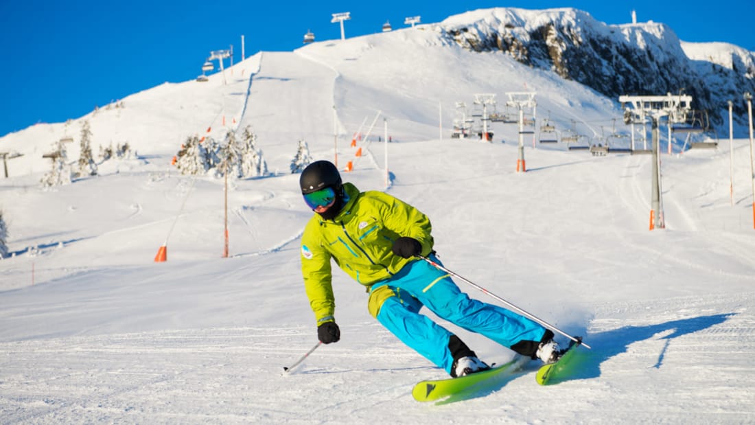 Person i fargerike klær kjører alpint i en snøkledd skibakke, med fjell og blå himmel i bakgrunn.