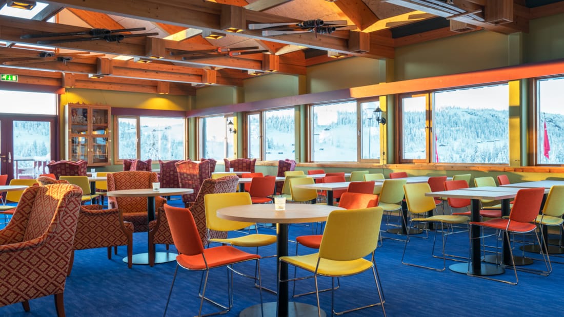 Salle à manger avec chaises colorées, grand tableau représentant Skeikampen au mur et grandes baies donnant sur le paysage à l’auberge de montagne Austlid