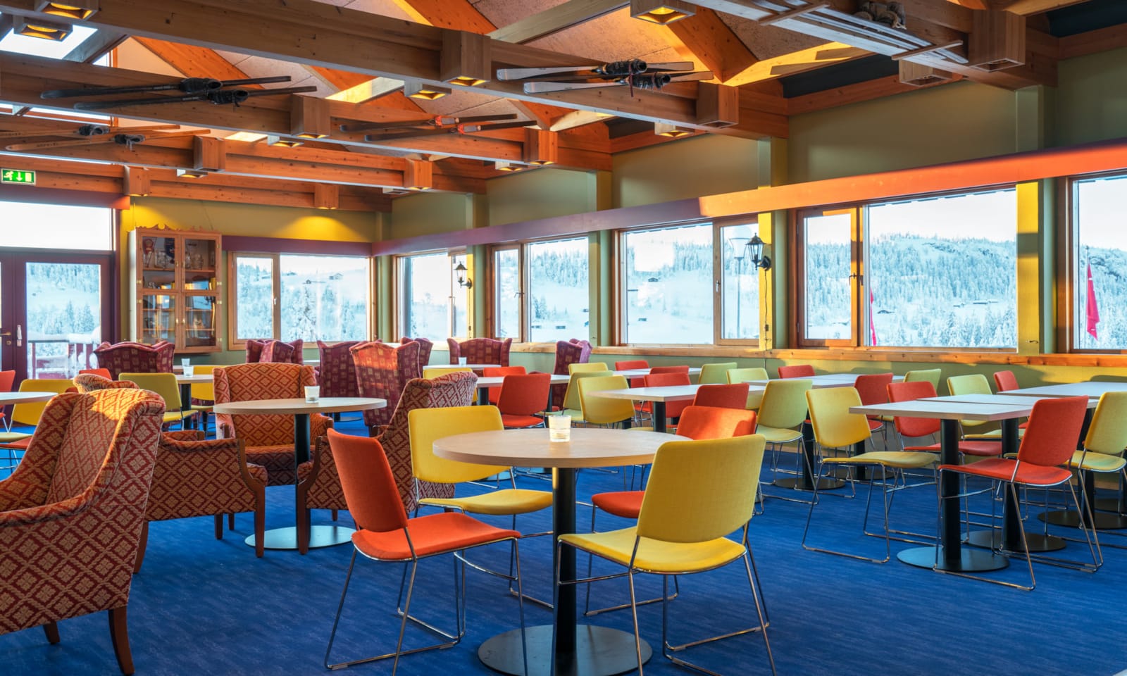 Dining area with colourful chairs, large painting of Skeikampen on the wall and large windows that look out onto the landscape of the Austlid fjellstue