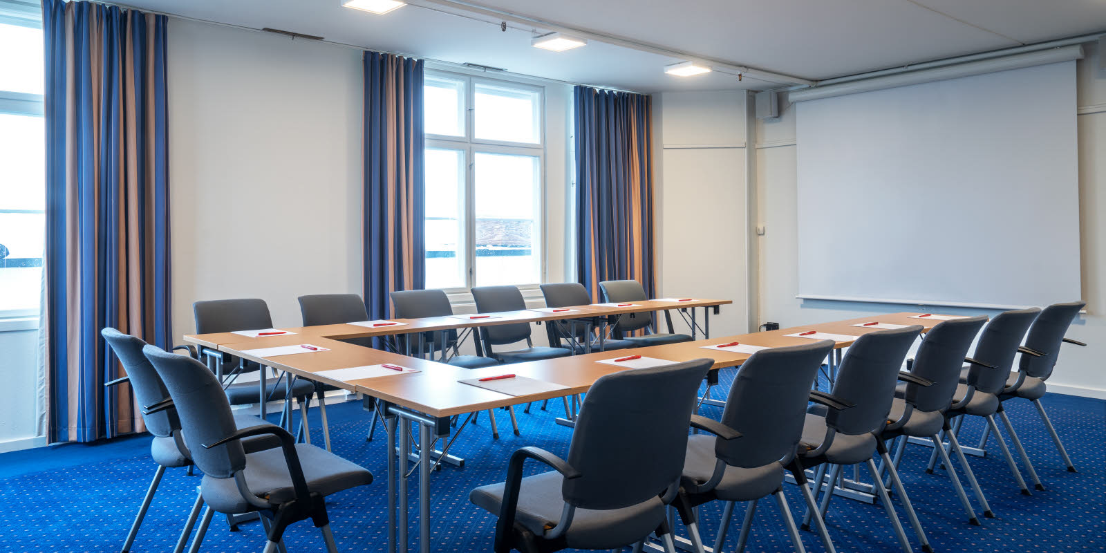 Prestkampen, u-bord med mørke stoler i lyst rom med flere vinduer, blått teppe og blå gardiner fra en annen vinkel