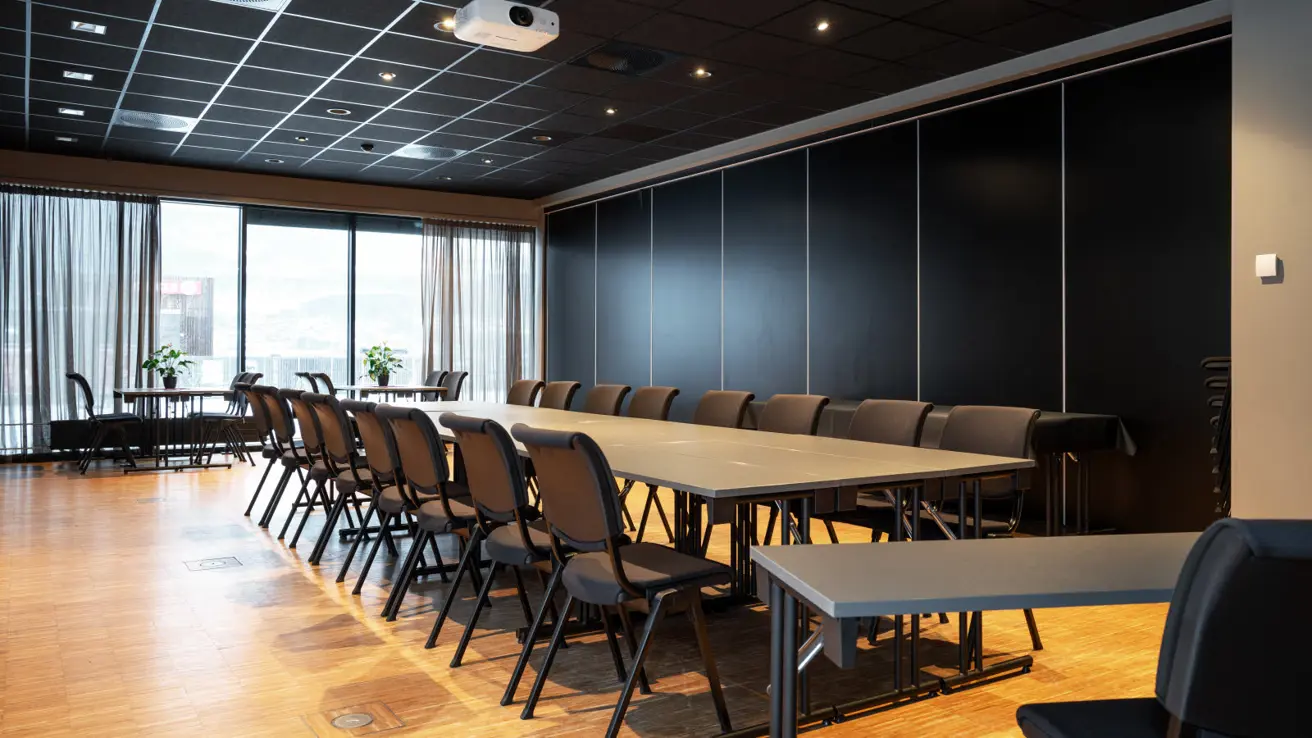 Vergaderruimte Solveig met lange tafel met 16 zitplaatsen. Op de achtergrond is de scheidingswand van de naastgelegen ruimte te zien. Op de voorgrond is een tafel met twee zitplaatsen, op de achtergrond twee tafels met vier zitplaatsen bij de ramen.