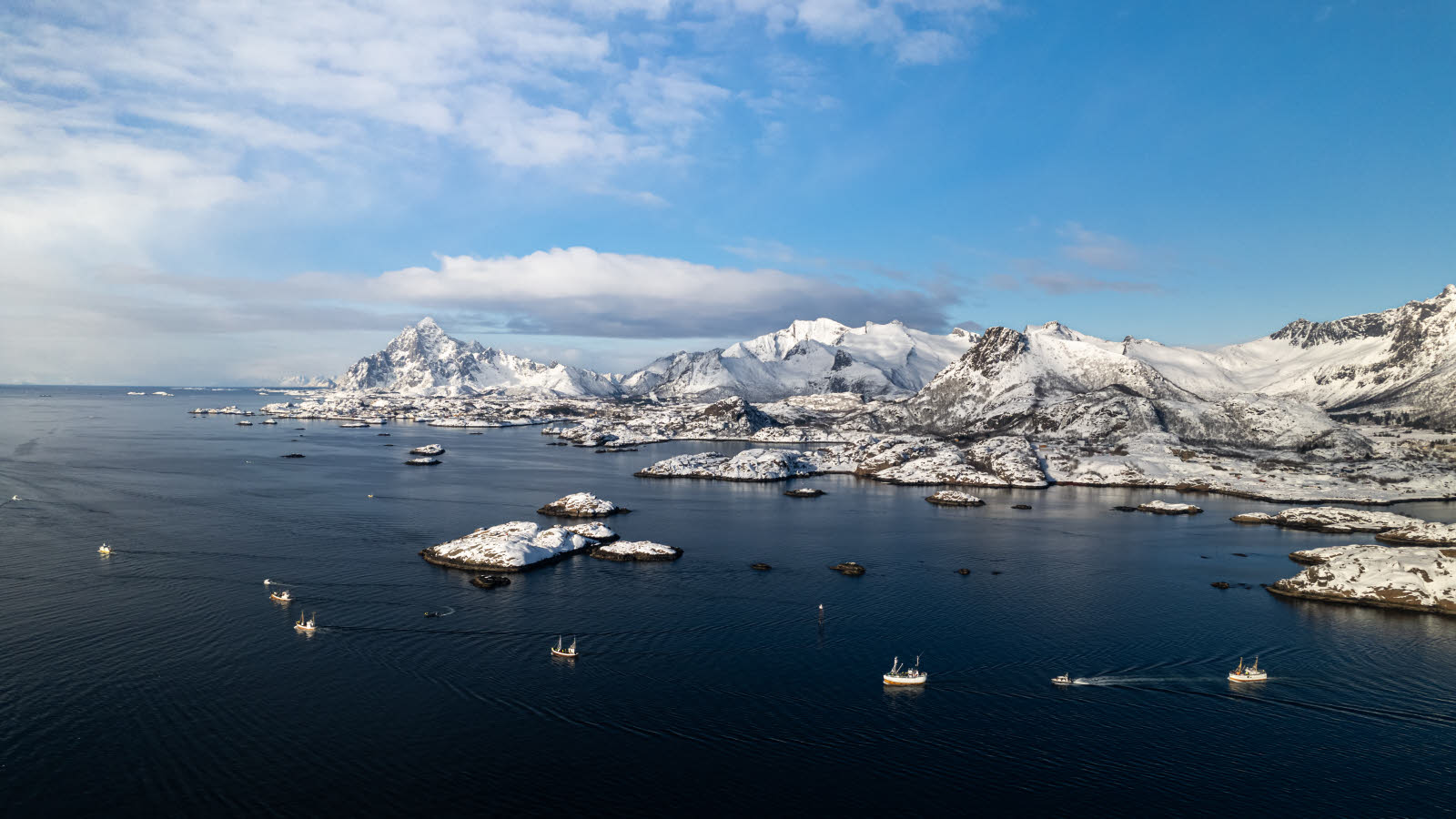 Lots of fishing boats on the water. Snow-covered mountains in the background.