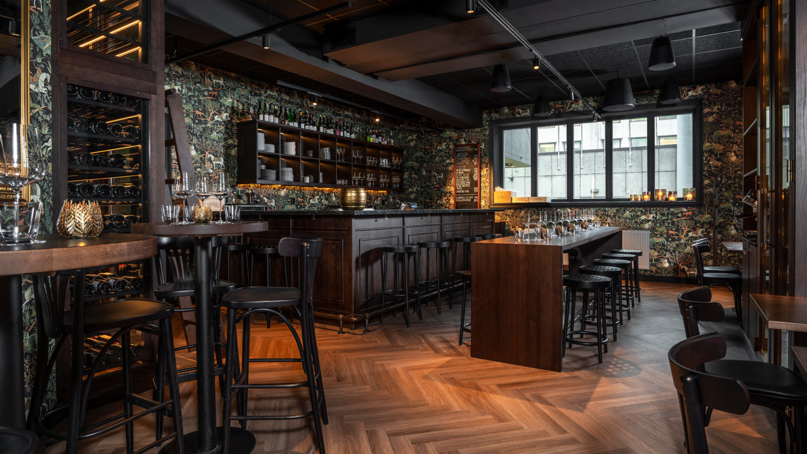 The Grotten beer and wine bar with a bar lined with round bar stools, dark wallpaper featuring trees, horses and other animals and plants, a long banquet table with matching dark stools, tall bar tables, large window and parquet floor.