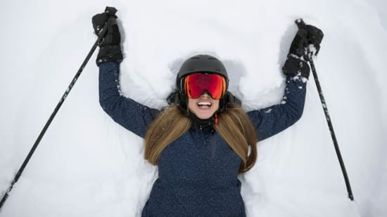 Kvinne som ligger med ryggen på snø og sportsbriller påmed skistaver i hendene og sportbriller på