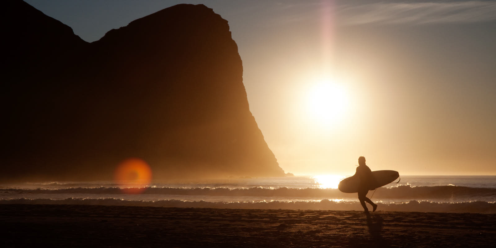 En surfer går ved vannkanten i solnedgang, med surfebrett under armen.