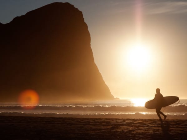 En surfer går ved vannkanten i solnedgang, med surfebrett under armen.