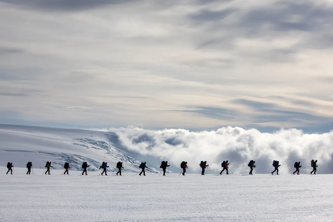 Lang rekke med mennesker som g&aring;r i taulag over en bree med ski og sekk p&aring; ryggen, masse sn&oslash; i forkant og bakkant p&aring; bildet.