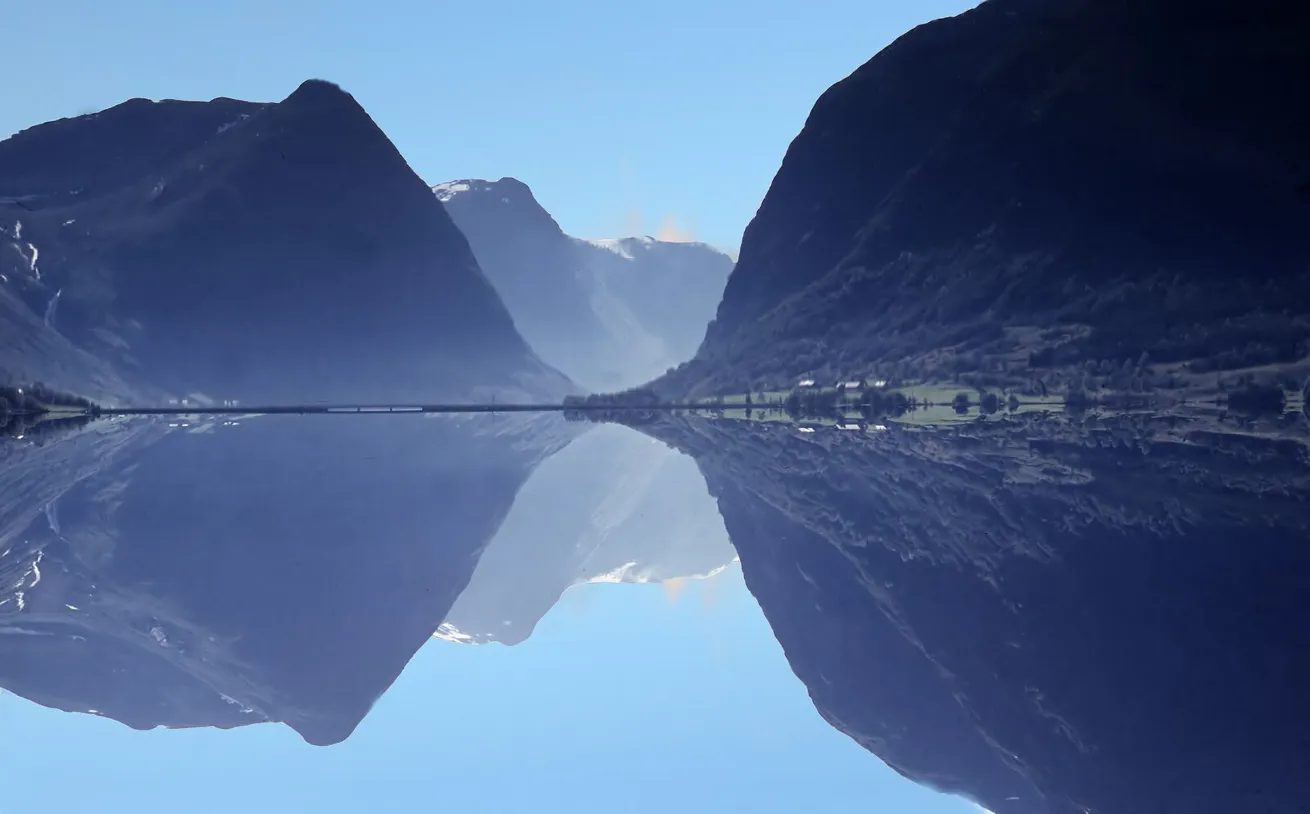 Bilde av to fjell som m&oslash;ter hverandre sett fra vannet. Flott speilbilde av fjellene i vannet med bl&aring; himmel bak.
