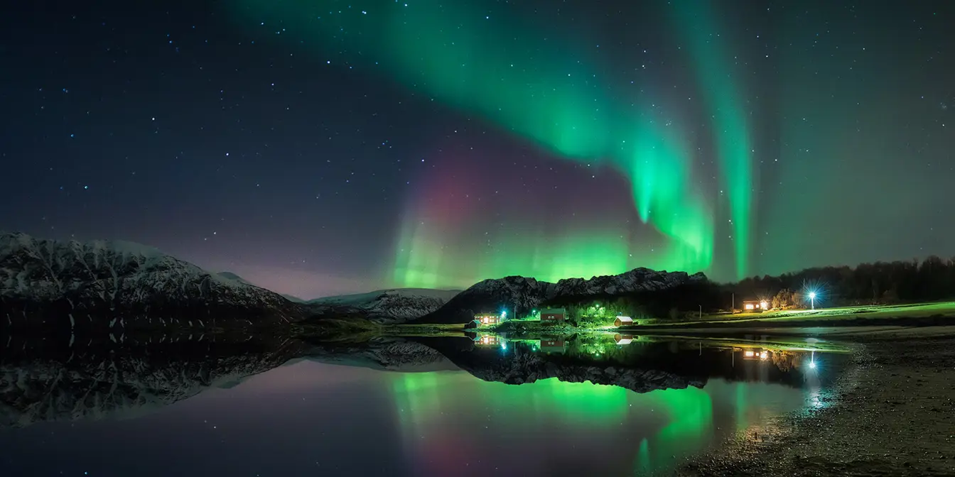 Türkis und violettes tanzendes Nordlicht, das das Meer mit Bergen und einer leuchtenden Stadt am Horizont reflektiert.