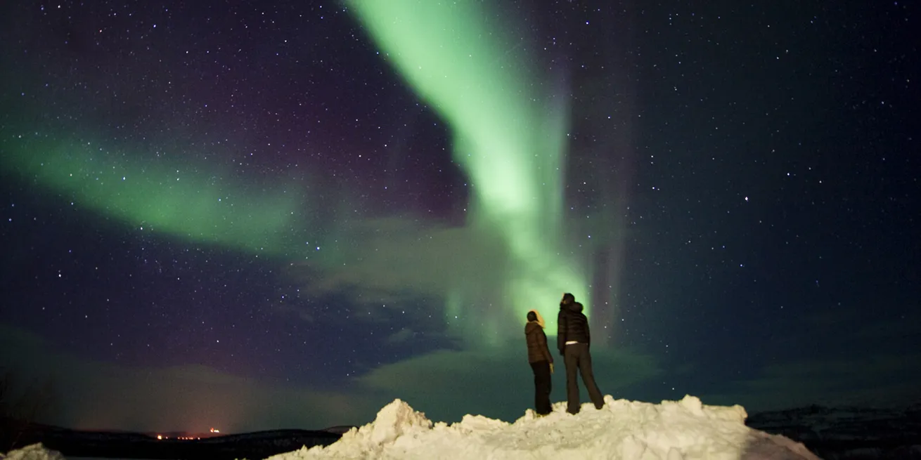 Deux personnes se tiennent sur un tas de neige et regardent les aurores boréales vertes dansant dans un ciel étoilé.