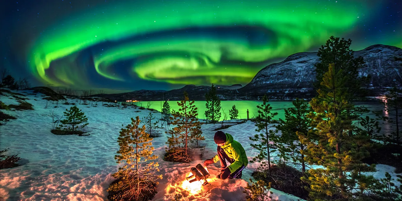 Personne assise sur ses genoux allumant un feu dans un paysage hivernal le long d’un lac avec des aurores boréales vertes tourbillonnantes dans le ciel.