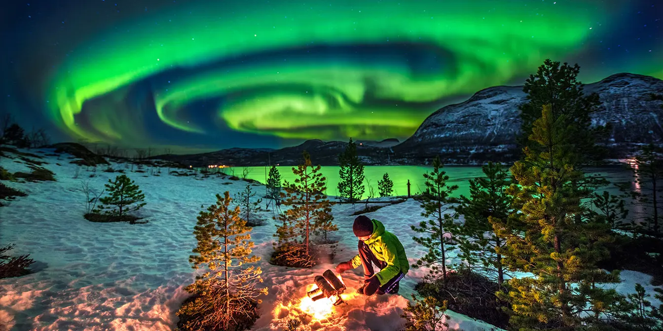 Eine Person sitzt auf der Hocke und entzündet ein Feuer in einer winterlichen Landschaft entlang eines Wassers mit grünem Nordlicht am Himmel.