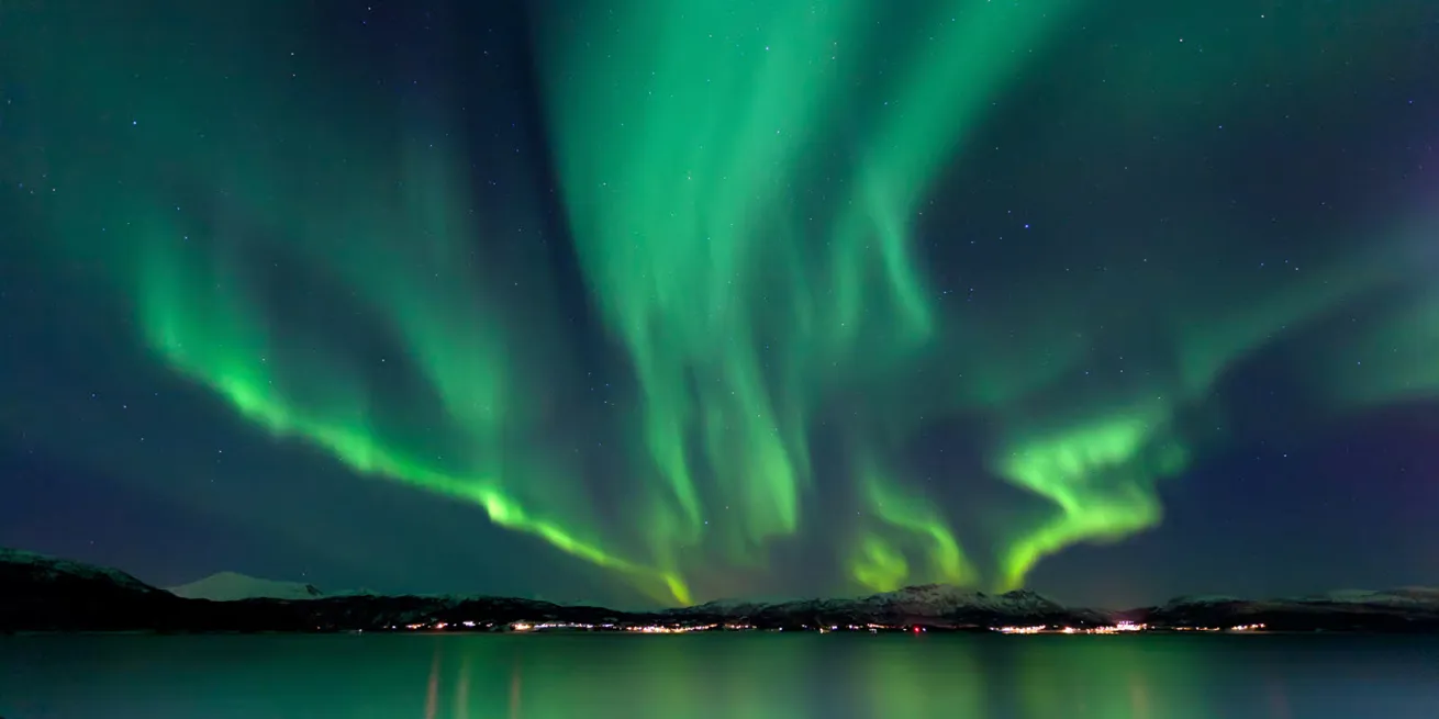 Aurore boréale verte dansant dans le ciel sur une chaîne de montagnes et lumières urbaines à l’horizon.