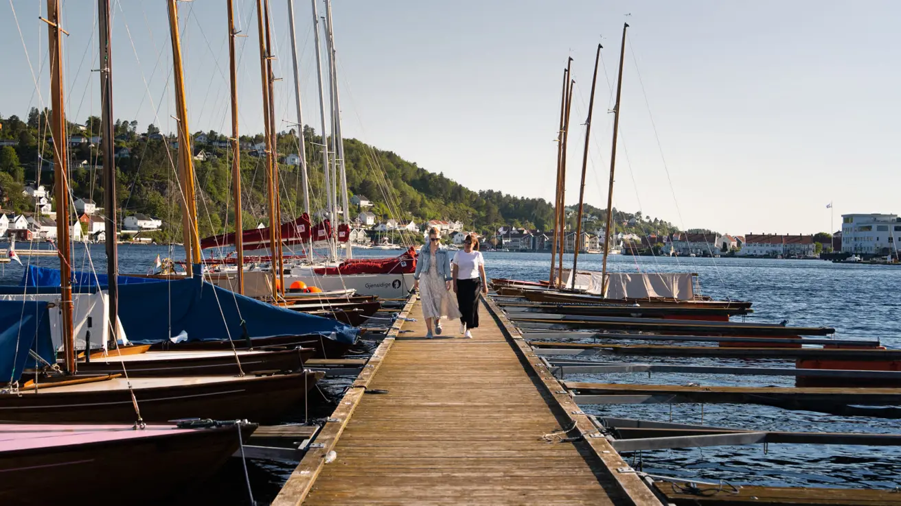 Two women strolling on a pier surrounded by boats.