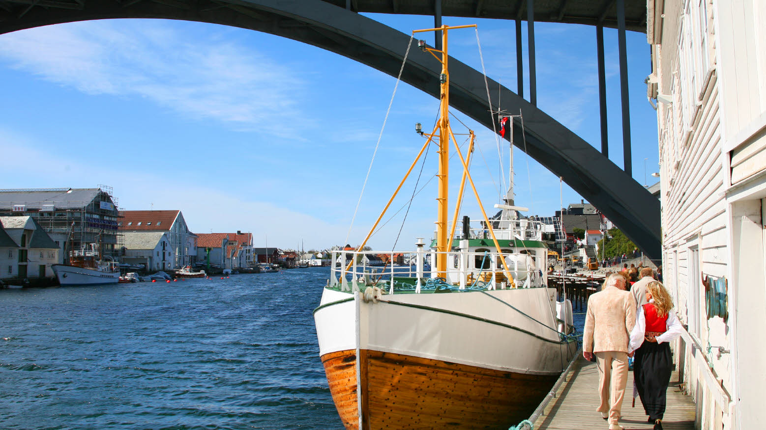 Kvinde og mand går under en bro med en båd fortøjet i vandet.