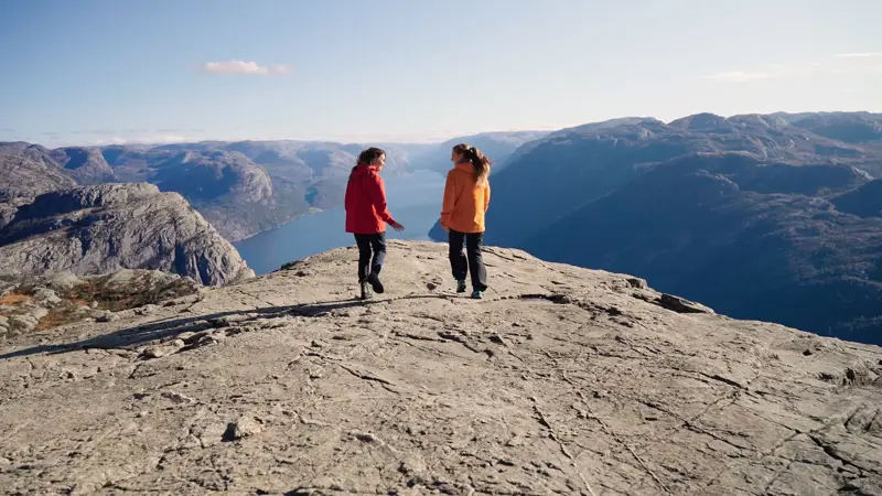 Twee vrouwen lopen naar de rand van Preikestolen. Op de achtergrond is de Lysefjord van bovenaf te zien, omringd door hoge bergen.