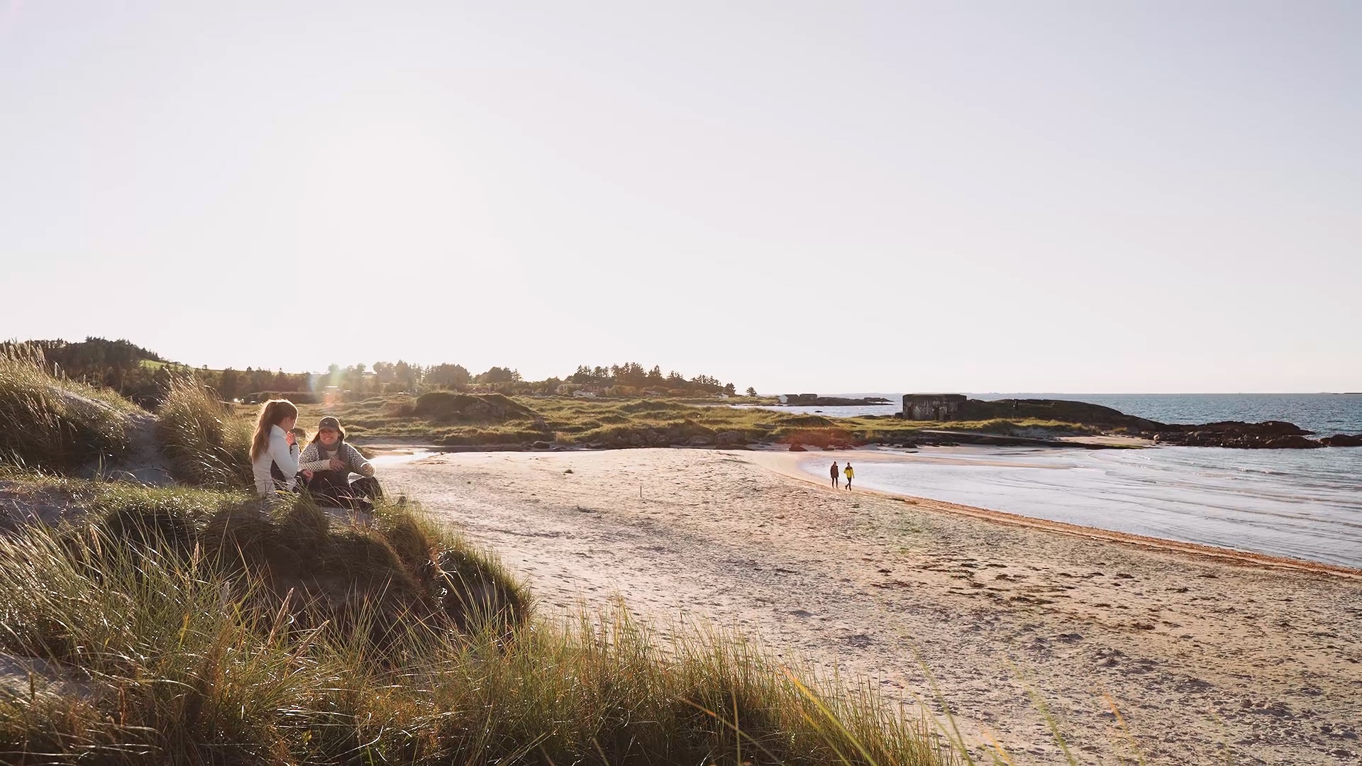 Lang sandstrand med sanddyner i forgrunnen og havet i bakgrunnen. Lett overskyet vær med solen som titter igjennom og lager fine farger i bildet. To mennesker kan skimtes på tur lengst bort i bildet.