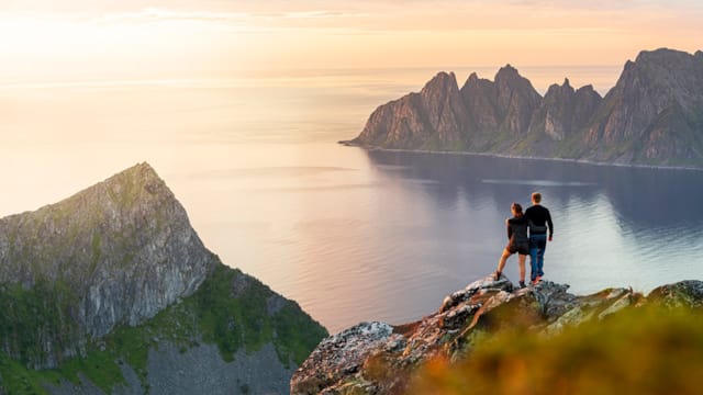 Two people standing on a mountain at sunset and looking out across the water and mountain landscape in Tromsø