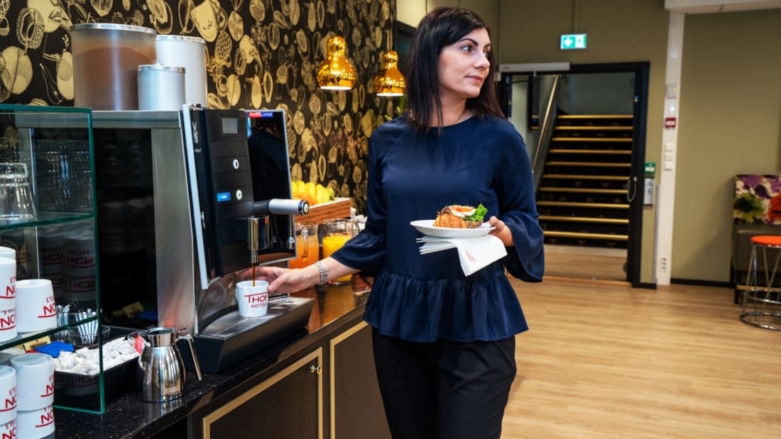 Kvinne som fyller kaffe fra en kaffemaskin, mens hun holder mat i en serviett i den andre hånden. Kvinnen befinner seg i et pauseområde på et konferansehotell.