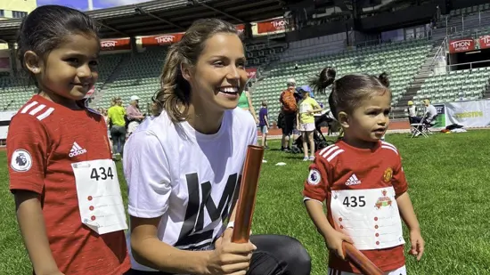 Smilende kvinne på huk med to barn i fotballdrakt stående på hver sin side. Friidrettsarena i bakgrunnen.