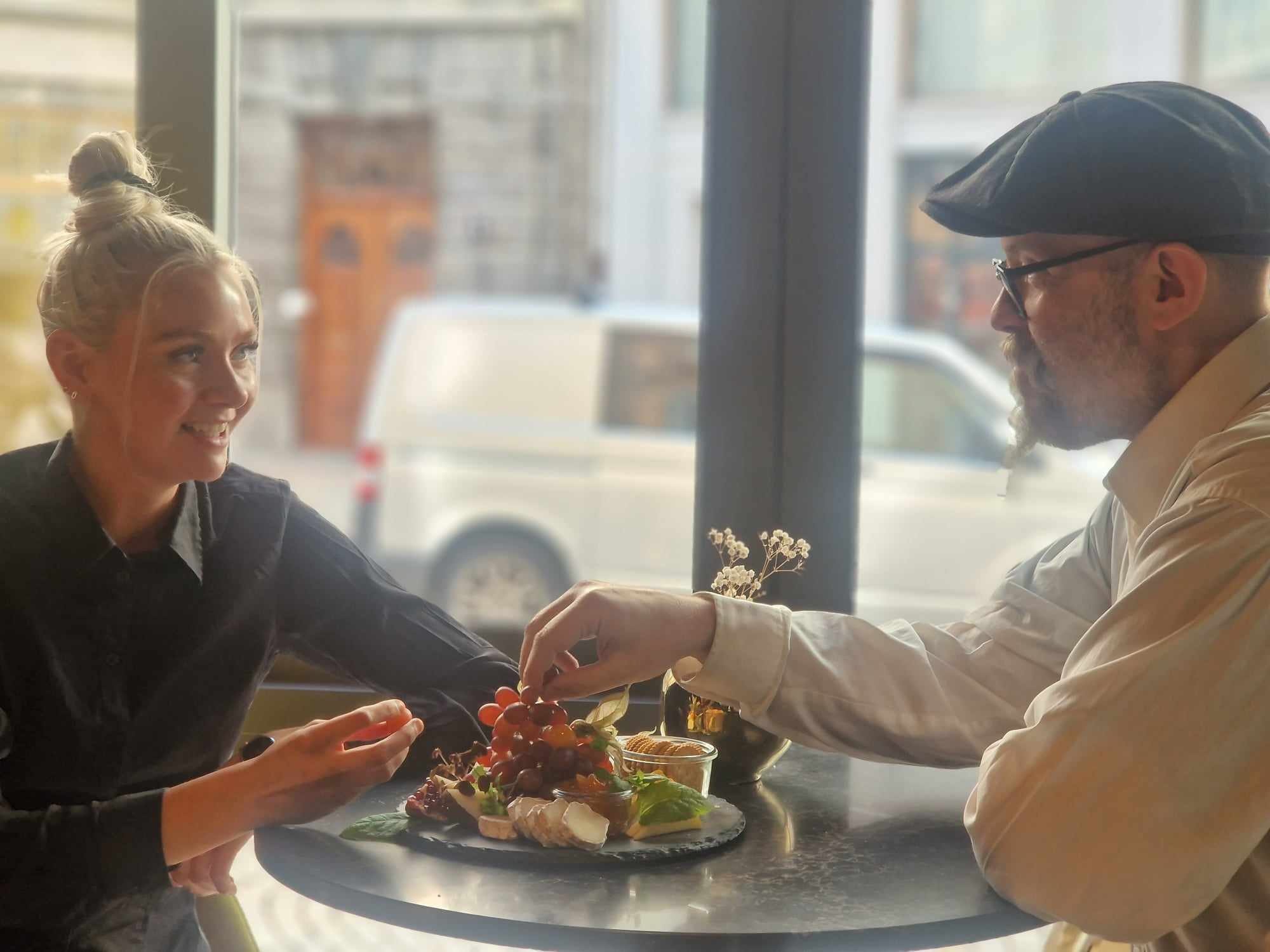 Two people sitting across from each other sharing a platter of cheese and fruit. Windows facing the street in the background. 