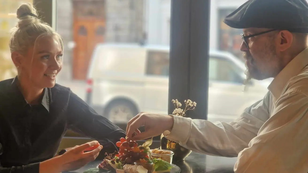 Two people sitting across from each other sharing a platter of cheese and fruit. Windows facing the street in the background.