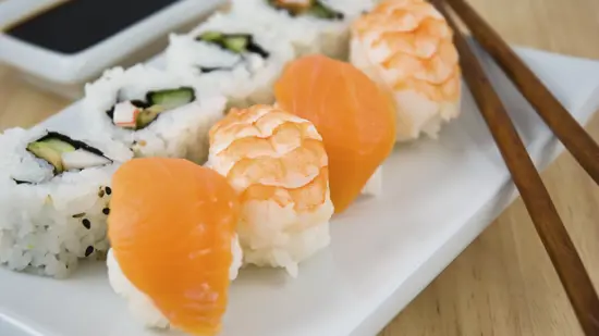 Sushi med laks og ris som ligger dandert på et hvit fat på et bord i eik med spisepinner i mørkt tre ved siden av sushien.